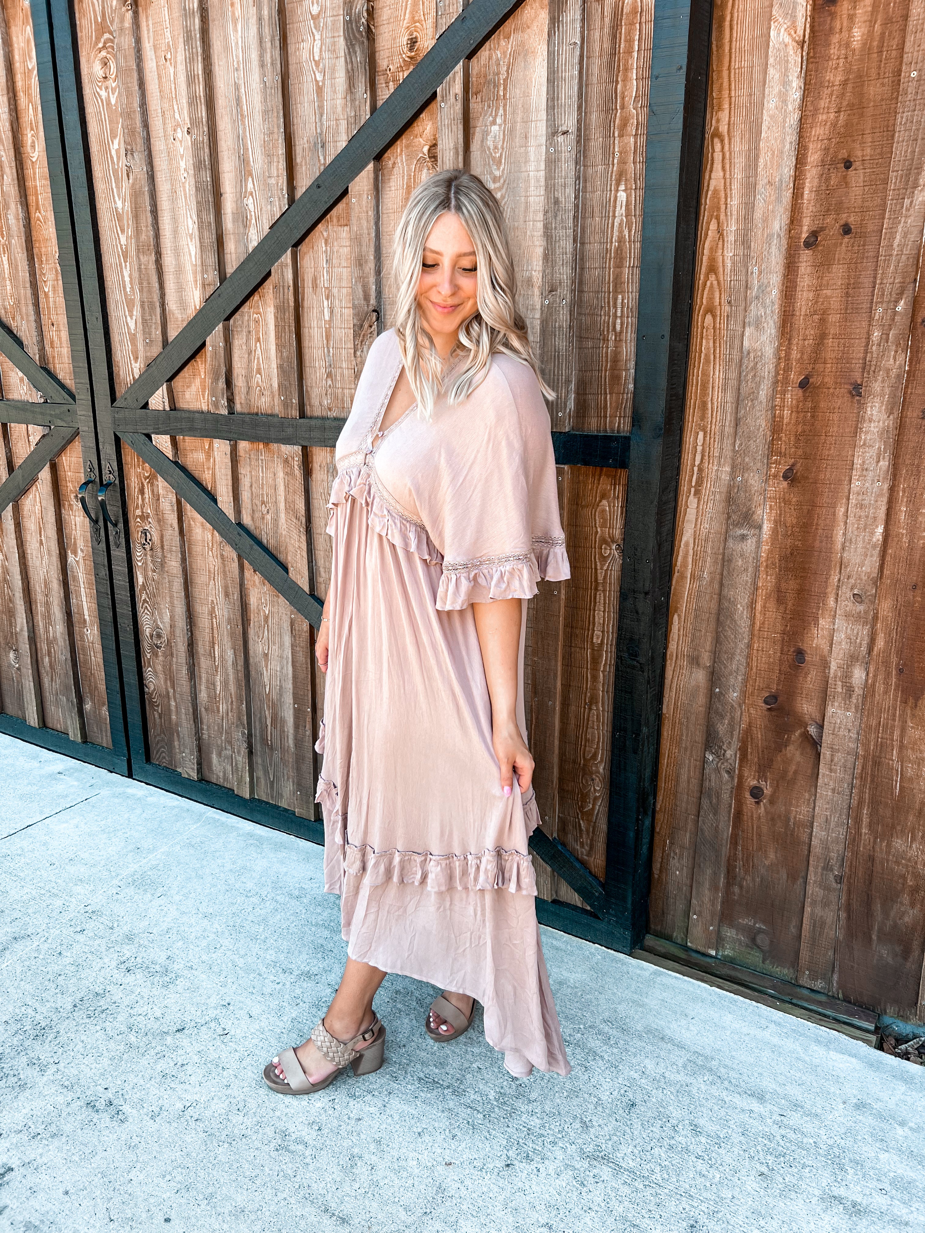 A boho maxi dress in a taupe color, featuring ruffle designs throughout enhanced with delicate lacy details along the ruffles. The dress has short, flowy sleeves and a V-neckline accented with small buttons at the top. The back mirrors the front with a V-neck as well, adding a harmonious and elegant detail. The combination of lace, ruffles, and the soft taupe shade lends the dress a romantic and vintage appeal, perfect for various occasions