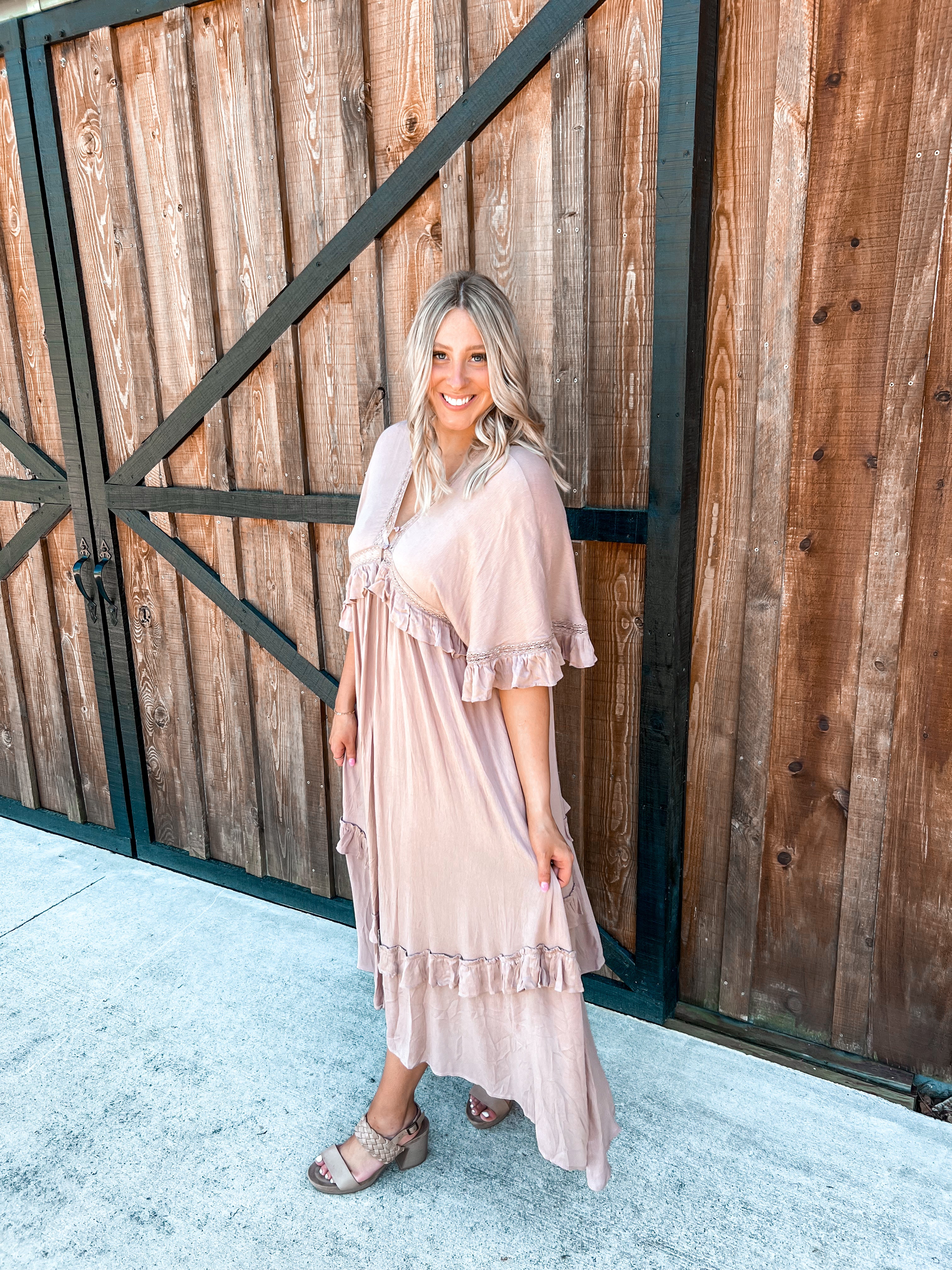 A boho maxi dress in a taupe color, featuring ruffle designs throughout enhanced with delicate lacy details along the ruffles. The dress has short, flowy sleeves and a V-neckline accented with small buttons at the top. The back mirrors the front with a V-neck as well, adding a harmonious and elegant detail. The combination of lace, ruffles, and the soft taupe shade lends the dress a romantic and vintage appeal, perfect for various occasions